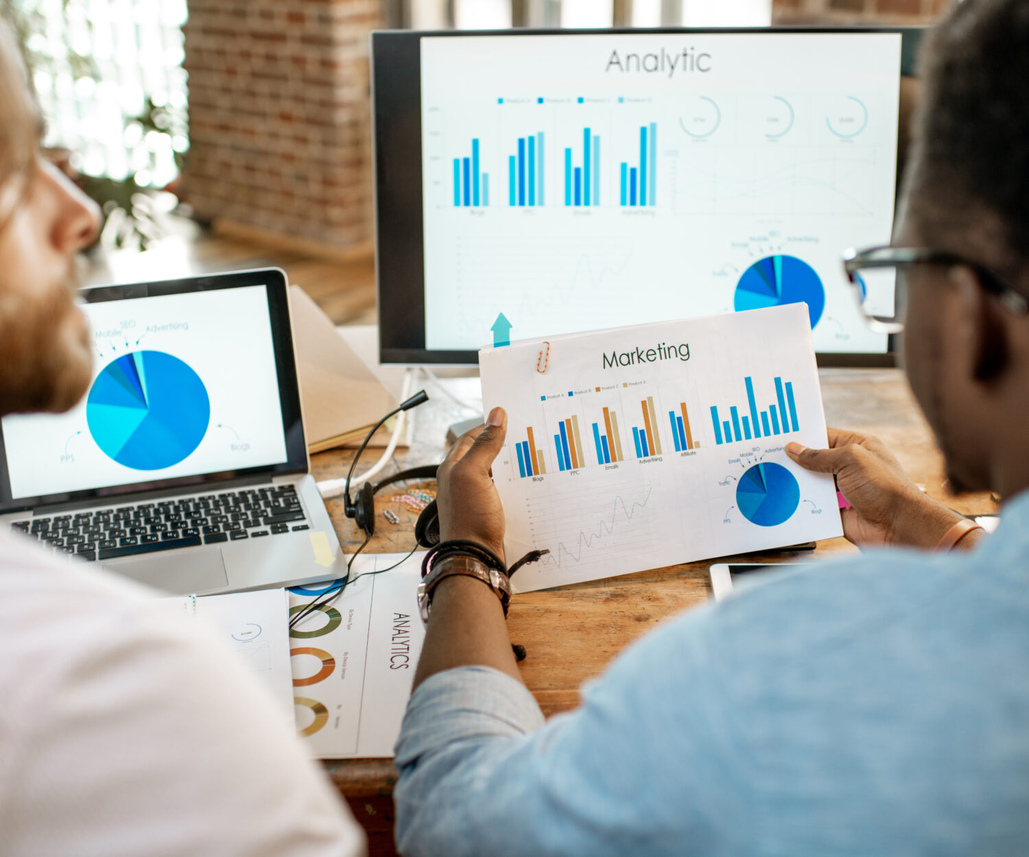Two male coworkers working with analitycs sitting together at the working place with computer, laptop and documents. View from behind focused on the documents